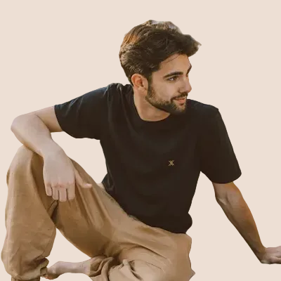 Man sitting on the ground wearing a black t-shirt and beige pants against a brown background